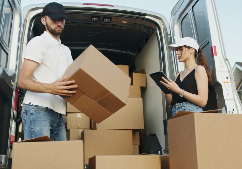 Two delivery workers handling cardboard boxes by a van, checking orders on tablet.